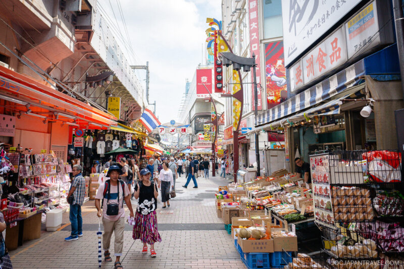 A busy outdoor market street in Japan, Ameyoko Market buzzes with people shopping, colorful Japanese signs, and stalls selling produce, snacks, and goods under a railway bridge.