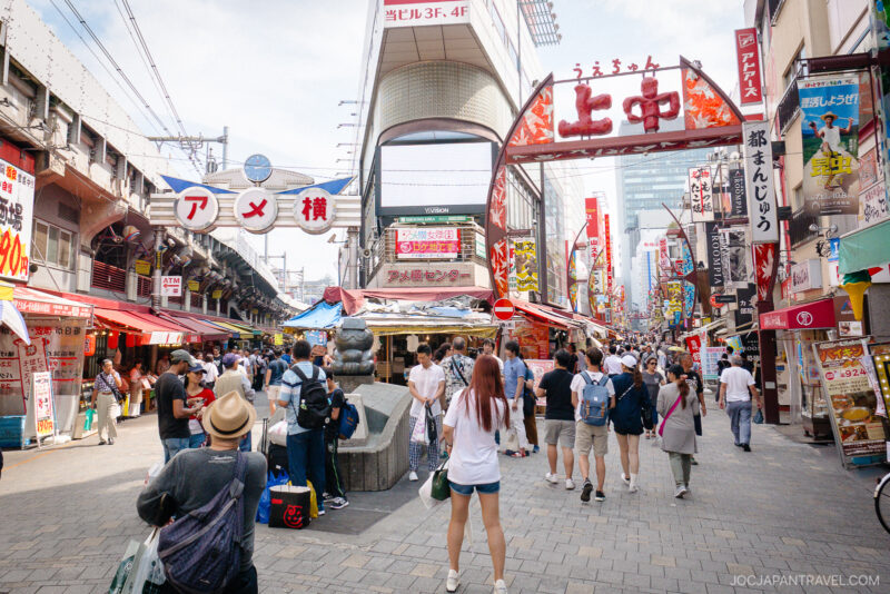 A busy pedestrian street market in Japan, Ameyoko Market is alive with people walking among colorful shops and food stalls under signs with Japanese characters. The area is lively and bustling with activity.