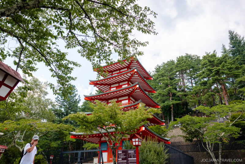 A red and white five-story pagoda in Arakurayama Sengen Park is surrounded by green trees, with a man in a white hat standing to the left and a partly cloudy sky above.
