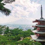 Arakurayama Sengen Park (新倉山浅間公園) – The Classic Mt. Fuji View A traditional five-story pagoda stands among green trees in Arakurayama Sengen Park, with a distant city view and hazy Mount Fuji partially visible through clouds in the background.