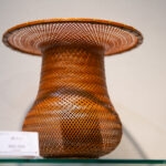 A finely crafted woven bamboo vase from the Beppu Bamboo Crafts Center, featuring a wide, flared rim and intricate zigzag patterns, is displayed on a glass shelf with a small price tag in front.