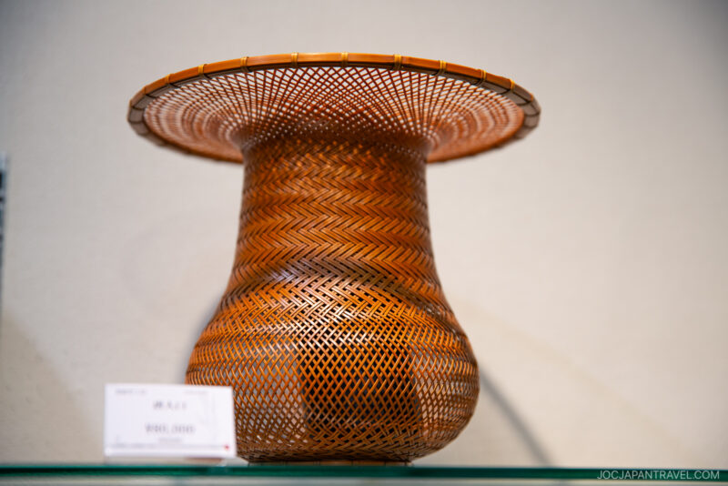 A finely crafted woven bamboo vase from the Beppu Bamboo Crafts Center, featuring a wide, flared rim and intricate zigzag patterns, is displayed on a glass shelf with a small price tag in front.