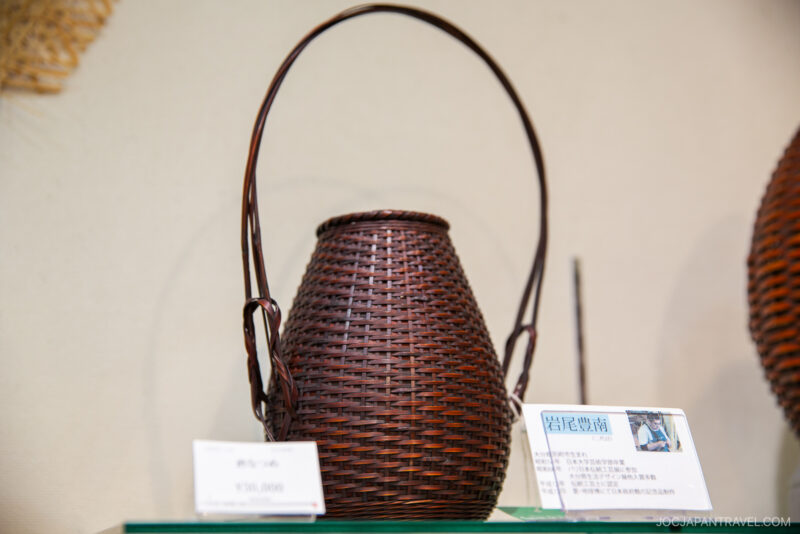 A handcrafted brown woven basket with a tall handle is displayed on a glass shelf at the Beppu Bamboo Crafts Center, accompanied by informational cards in Japanese.