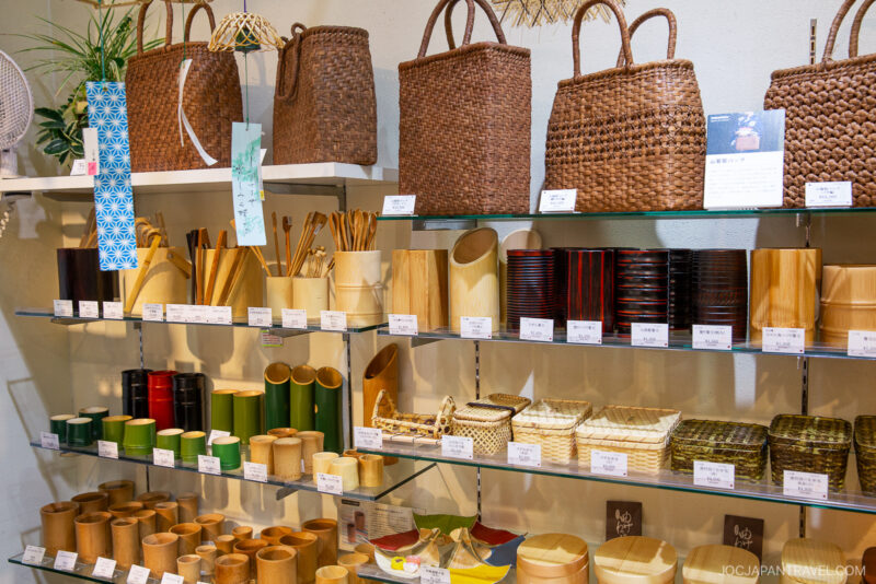 Shelves at the Beppu Bamboo Crafts Center display various bamboo and woven goods—baskets, cups, bowls, trays, and chopsticks—neatly organized with price tags and informational cards beside each item.