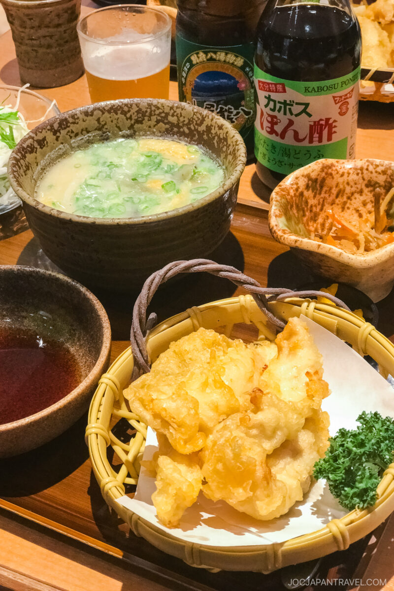 A Japanese meal featuring Beppu Chicken Tempura Toriten in a basket, a bowl of soup with green onions, dipping sauces, ponzu sauce, and side dishes on a wooden table.