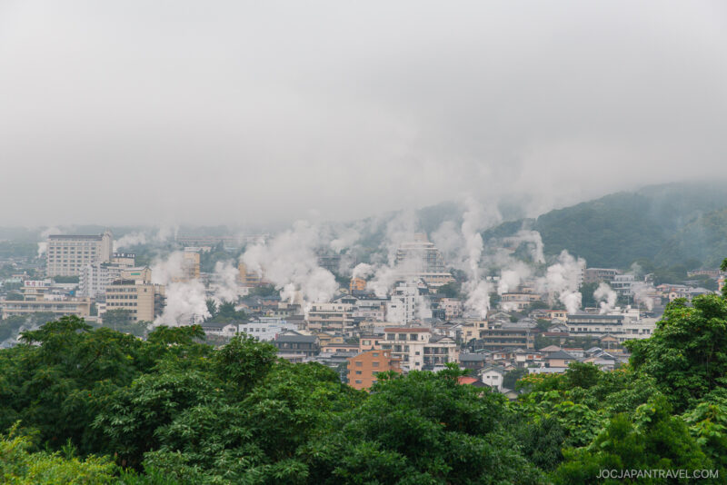 A cityscape with numerous buildings emitting white steam, surrounded by green hills and partially obscured by fog or low clouds—a scene straight from a Beppu Onsen Travel Guide. Dense greenery fills the foreground.