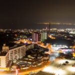 A cityscape at night with brightly lit buildings and streets, distant lights twinkling along the coastline, and the iconic Beppu Suginoi Hotel adding to the vibrant urban scene against the dark sky.