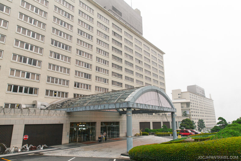 A large, modern multi-story hotel building, resembling the Beppu Suginoi Hotel, features a glass-covered entrance, white exterior, and numerous windows, set on a cloudy day with greenery and neatly trimmed bushes in the foreground.