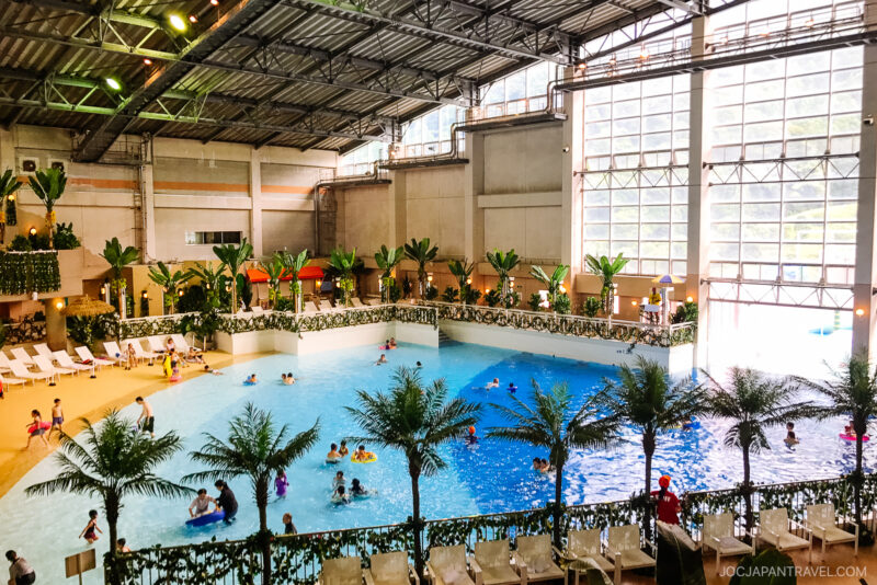 Indoor water park at Beppu Suginoi Hotel features a large wave pool, artificial palm trees, and lounge chairs. Several people swim and play under a high glass ceiling with natural light streaming in.