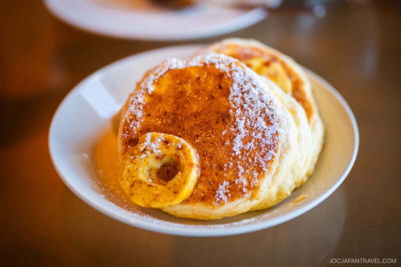 A plate of fluffy pancakes dusted with powdered sugar and garnished with banana, served on a white dish at Bills Odaiba at DECKS.