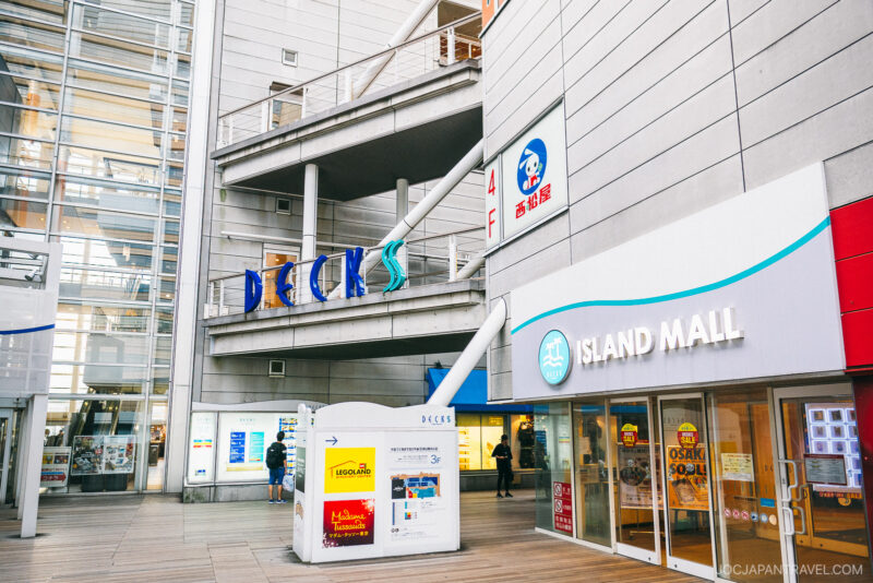 Modern shopping mall exterior with signs for DECKS Tokyo Beach and ISLAND MALL, glass walls, and people walking near a directory board. The setting is bright, clean, and inviting with multiple floors visible.