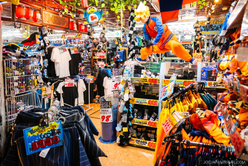 Colorful Akihabara Don Quijote store interior filled with clothes, accessories, sale signs, and a large Dragon Ball Goku figure suspended above the aisles. Bright lights and dense displays create a lively, busy atmosphere.