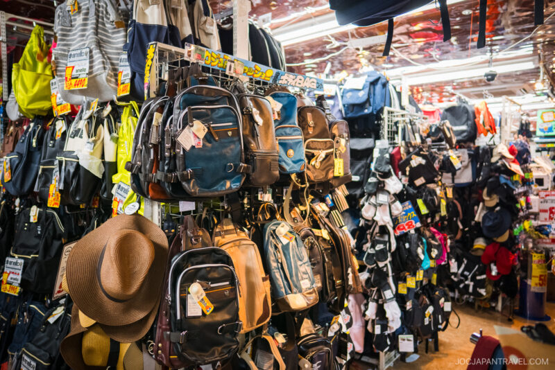 A store display at Akihabara Don Quijote features a variety of backpacks, hats, and bags, all closely packed with price tags. The shop is busy and colorful, with products hanging on shelves and racks.