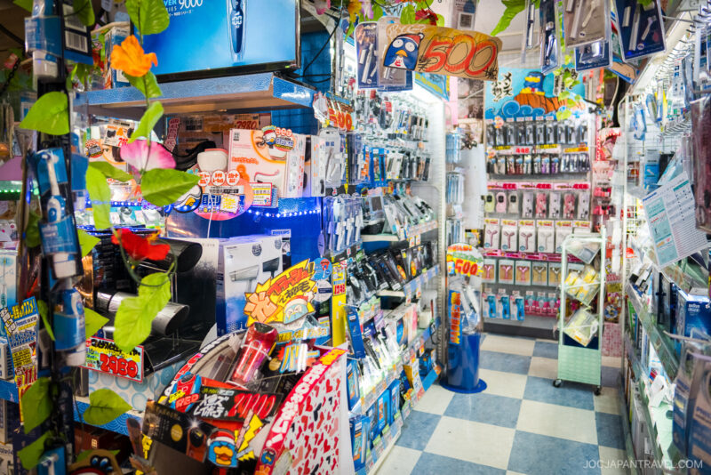 A brightly lit aisle in Akihabara Don Quijote, packed with electric shavers, razors, and grooming products. Colorful price tags and packaging fill the shelves; fake green vines hang as decoration.
