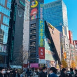 A busy street in Tokyo’s Akihabara with crowds of people, many wearing masks, walking among tall buildings covered in colorful signs and ads, including a large Don Quijote store with vibrant anime-themed signage.