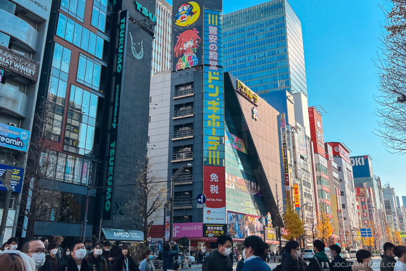 A busy street in Tokyo’s Akihabara with crowds of people, many wearing masks, walking among tall buildings covered in colorful signs and ads, including a large Don Quijote store with vibrant anime-themed signage.