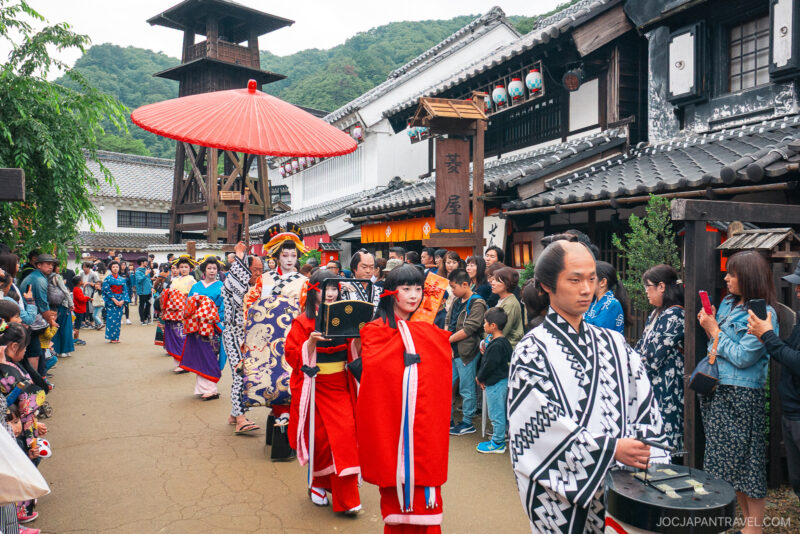 A traditional Japanese parade at Edo Wonderland Nikko Edomura features people in colorful kimonos and costumes walking down a street lined with old-style buildings, as onlookers watch and take pictures. A large red umbrella is held above one participant.
