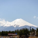 Exploring Fuji-Q Highland (富士急ハイランド) – Thrills at the Foot of Mt. Fuji Snow-capped Mount Fuji rises under a clear blue sky, with the Fuji-Q Highland roller coaster on the left, trees at the base, and a small gas station and parked cars in the foreground.