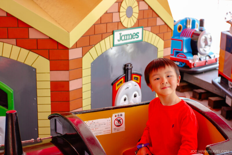 A young boy in a red shirt smiles while sitting inside a ride at Fuji-Q Highland, with a colorful Thomas & Friends-themed backdrop featuring train faces and a building labeled James behind him.