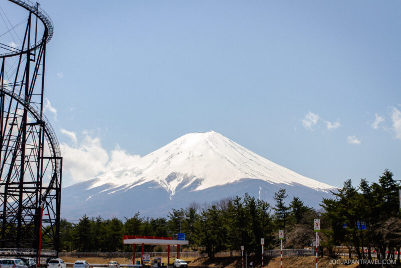 Snow-capped Mount Fuji rises under a clear blue sky, with the Fuji-Q Highland roller coaster on the left, trees at the base, and a small gas station and parked cars in the foreground.