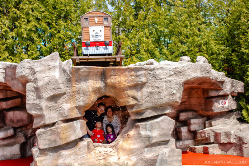 A family poses inside a rocky cave structure at Fuji-Q Highland theme park, with a character train resembling Troublesome Trucks from Thomas & Friends positioned on top. Green trees fill the background.