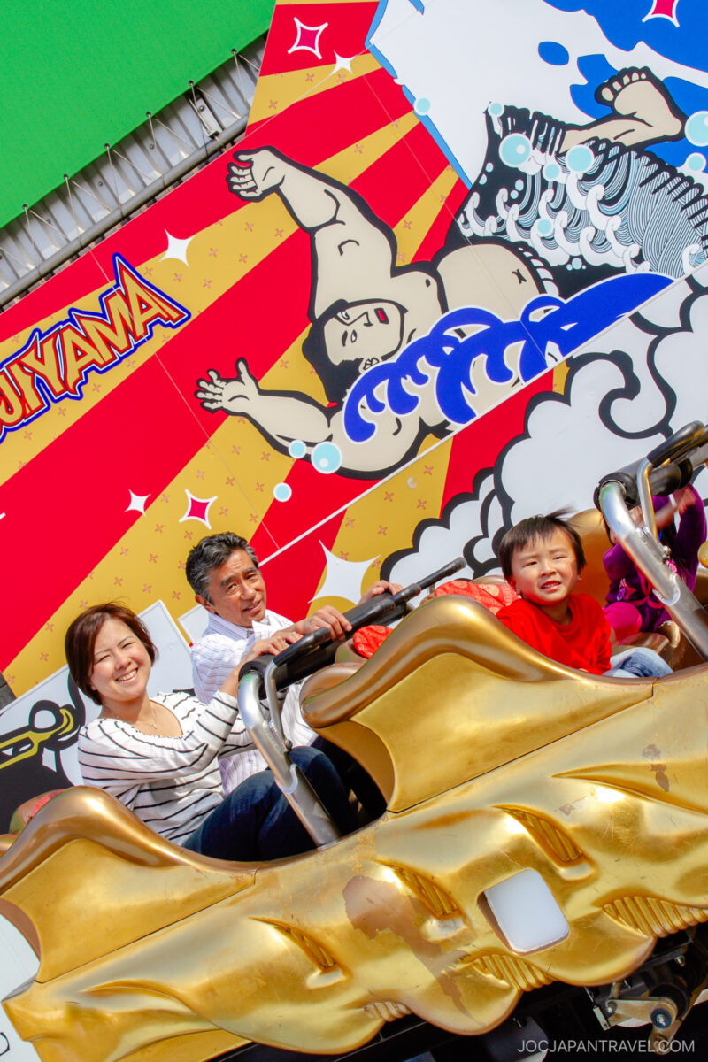 A smiling family of three rides a gold roller coaster at Fuji-Q Highland, set against a colorful backdrop featuring a sumo wrestler and bold patterns at the amusement park.