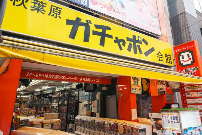 Street view of the brightly colored Akihabara Gachapon Hall, a Japanese capsule toy store with a yellow sign and rows of toy vending machines at the entrance. The shop’s vibrant interior and signage are clearly visible.