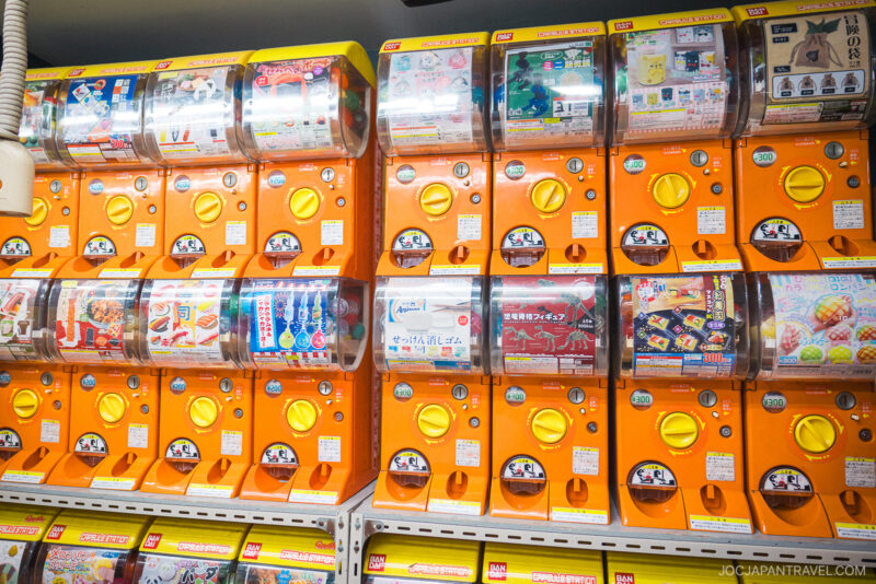 Rows of orange Japanese capsule toy machines (gashapon) at Akihabara Gachapon Hall display colorful toys and prizes in clear windows above yellow coin slots and handles.