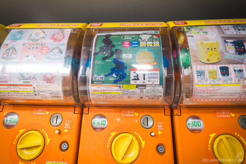 Three bright orange Japanese capsule toy vending machines (gashapon) from Akihabara Gachapon Hall, featuring animal and character figures. Each machine costs 300 yen per capsule.