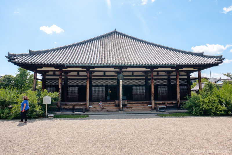 A traditional Japanese temple, Nara Gango-ji, with a tiled roof, wooden pillars, and open veranda, is surrounded by greenery. A person stands to the left, facing the building under a bright blue sky with scattered clouds.