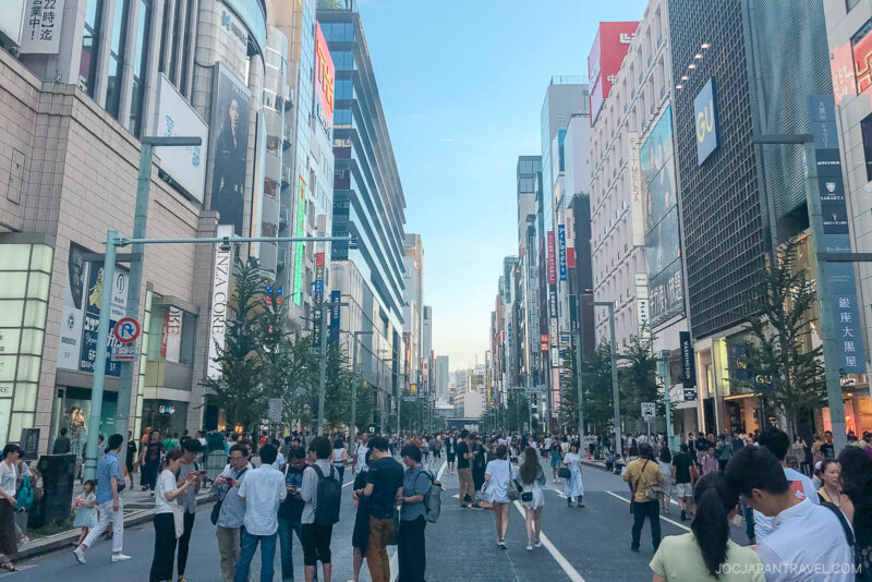 A busy city street lined with tall buildings and shops, filled with people walking and gathering under a clear sky. The road is closed to vehicles, transforming the area into a lively Ginza Hokosha Tengoku pedestrian zone.