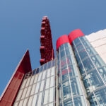 A low-angle view of a modern building with glass and red architectural elements, featuring part of the iconic HEP FIVE Ferris Wheel visible against a clear blue sky in the background.