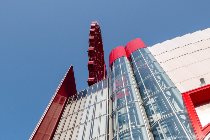 A low-angle view of a modern building with glass and red architectural elements, featuring part of the iconic HEP FIVE Ferris Wheel visible against a clear blue sky in the background.
