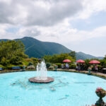 Hakone Gora Park (箱根強羅公園) – Gardens, Crafts, and Tea in Hakone A round fountain in a clear blue pool is surrounded by flowers and benches, with red umbrellas shading visitors at Hakone Gora Park. Green mountains and a partly cloudy sky complete the picturesque scene in the background.
