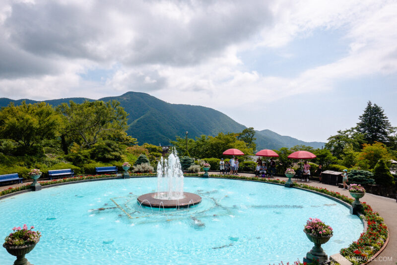 A round fountain in a clear blue pool is surrounded by flowers and benches, with red umbrellas shading visitors at Hakone Gora Park. Green mountains and a partly cloudy sky complete the picturesque scene in the background.