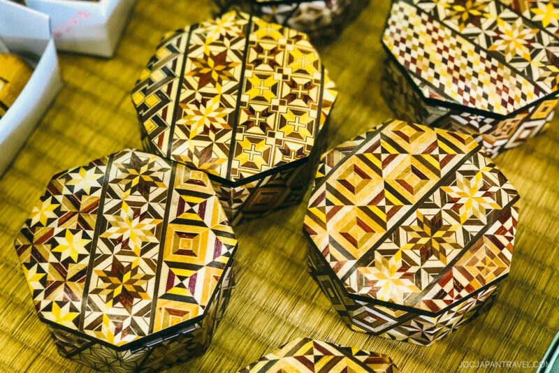 Several hexagonal wooden boxes, reminiscent of Hakone Himitsu-Bako, display intricate geometric marquetry patterns in shades of brown and yellow. Stars, triangles, and diamonds adorn their surfaces as they rest on a woven mat.