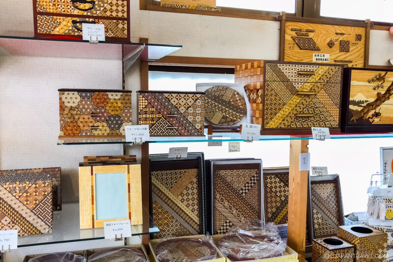 Shelves display traditional Japanese Yosegi marquetry boxes, trays, Hakone Himitsu-Bako puzzles with intricate geometric wood patterns in various colors and shapes. Price tags and small signs are visible near each item.