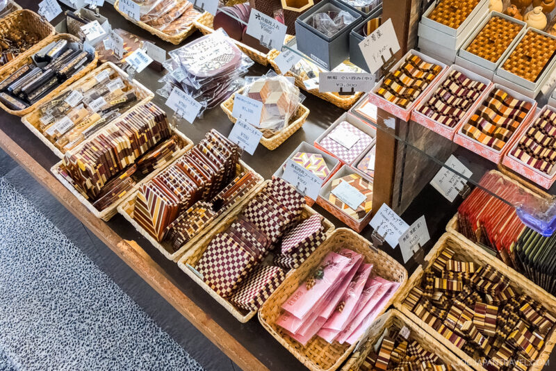 A display of assorted Japanese sweets, some nestled in Hakone Himitsu-Bako-inspired boxes, with handwritten labels in Japanese. The candy wrappers are colorful and patterned, adding to the inviting and vibrant presentation.