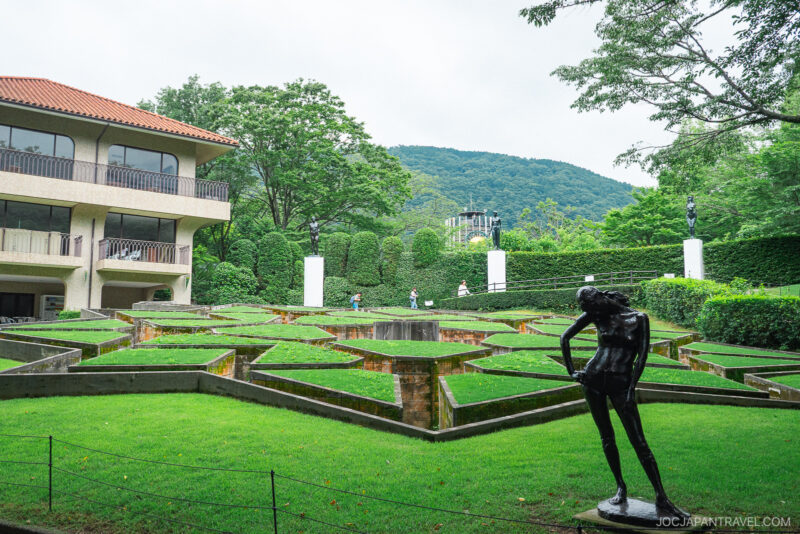 Sculpture garden with geometric grass shapes, a bronze statue in the foreground, and a modern building on the left, set against green hills at the Hakone Open-Air Museum.