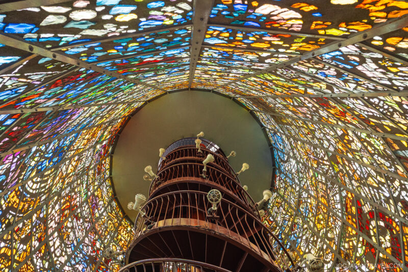 A spiral staircase ascends inside a tall, cylindrical tower at the Hakone Open-Air Museum, where colorful, intricate stained glass panels create a vibrant, mosaic-like effect as light streams through.