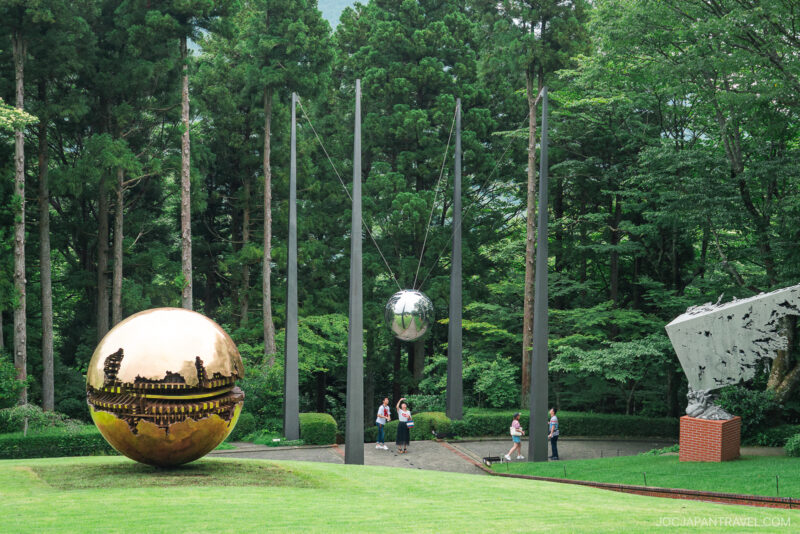 Sculptures of reflective silver and gold spheres stand amid tall trees and grass at the Hakone Open-Air Museum, while several people walk nearby in an outdoor park surrounded by greenery.