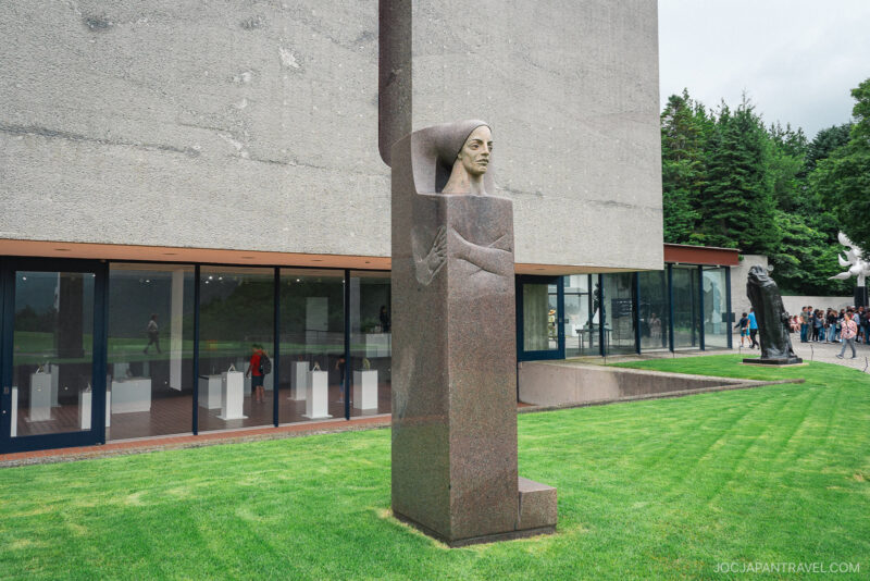 A tall stone sculpture featuring a woman carved into a column stands on a manicured lawn at the Hakone Open-Air Museum, set before a modern building with large windows; several other sculptures and visitors are visible in the background.