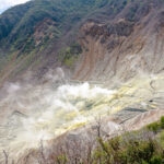 Hakone Ropeway & Owakudani Valley (大涌谷) – A Volcanic Adventure with Mount Fuji Views A volcanic valley near the Hakone Ropeway, with white steam rising from fumaroles, yellow sulfur deposits on rocky terrain, and green vegetation on surrounding slopes under a partly cloudy sky.