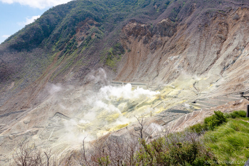 A volcanic valley near the Hakone Ropeway, with white steam rising from fumaroles, yellow sulfur deposits on rocky terrain, and green vegetation on surrounding slopes under a partly cloudy sky.