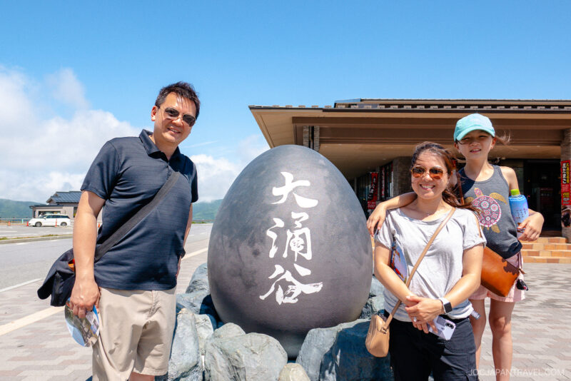 Three people stand smiling beside a large black egg-shaped stone with white Japanese characters at Owakudani Valley, near the Hakone Ropeway. They are outdoors on a sunny day, with mountains and a modern building in the background.