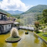 A picturesque garden at the Hakone Venetian Glass Museum features a pond, small boat, stone buildings, manicured greenery, a white bridge, and tall wire sculpture set against lush mountains under a partly cloudy sky.