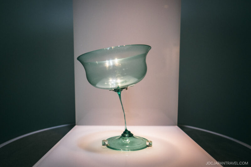 A delicate, green-tinted glass goblet with an asymmetrical, tilted bowl and thin stem is displayed on a white pedestal under focused lighting at the Hakone Venetian Glass Museum.