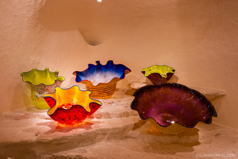 Colorful, wavy-edged glass bowls in red, yellow, green, blue, and purple are displayed on white stone steps against a textured beige wall at the Hakone Venetian Glass Museum. Light highlights their vibrant colors and unique shapes.