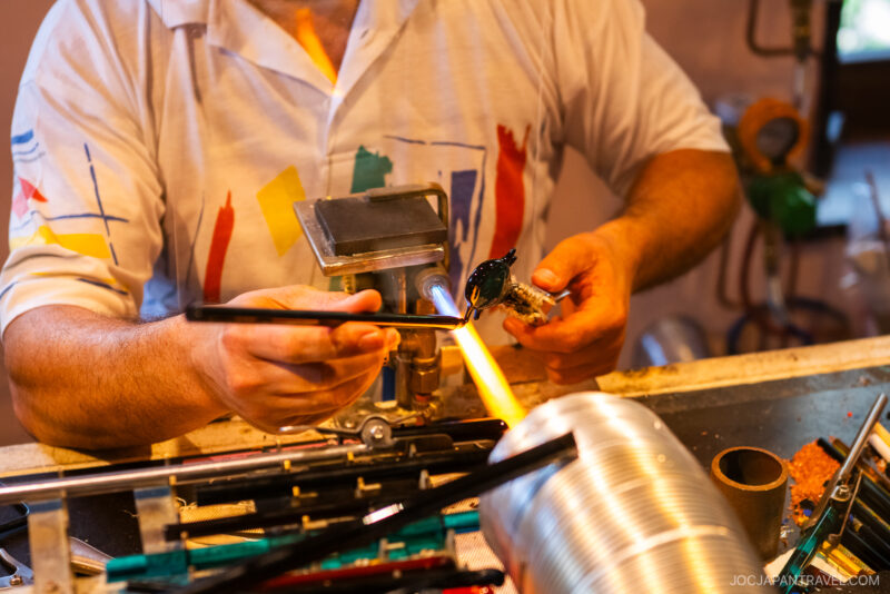 In a workshop at the Hakone Venetian Glass Museum, a person shapes a piece of glass with a torch, holding it with tongs while heating it. Colorful tools and equipment are visible on the workbench.