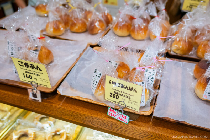 Various types of sweet buns from Hakone Watanabe Bakery are wrapped in clear plastic and displayed on trays, each with a Japanese price tag and description card in front.